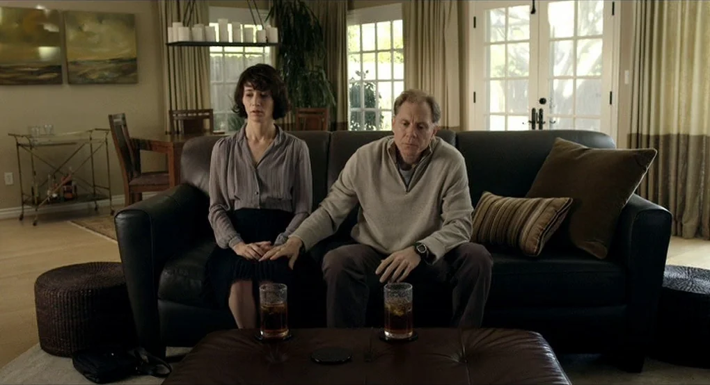 The Future (2011) movie still: medium — A furnished living room with a black leather sofa and matching ottoman. A man sits next to a woman and…