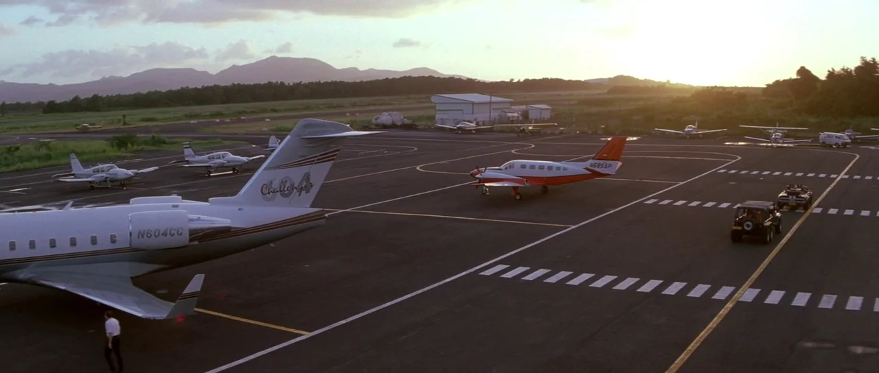 The Thomas Crown Affair (1999) movie still: establishing — An airfield at sunset features several light aircraft parked on a marked black tarmac.…