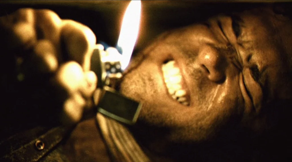 Buried (2010) movie still: closeup — A man lies in a dark, confined space illuminated only by a small flame. The man grimaces and squints his eyes…