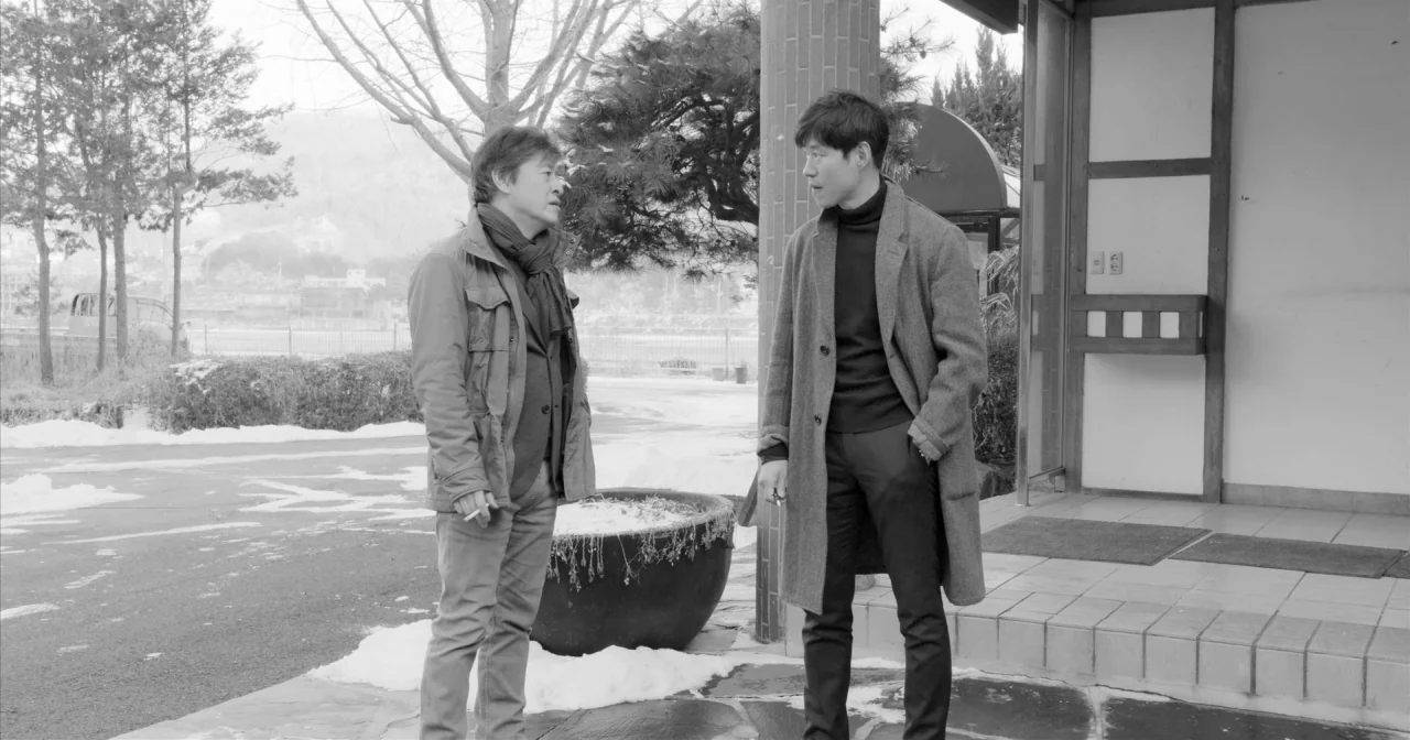 Hotel By The River movie still: fullbody — An outdoor area beside a building featuring stone tiling and a large concrete planter. Two men stand…
