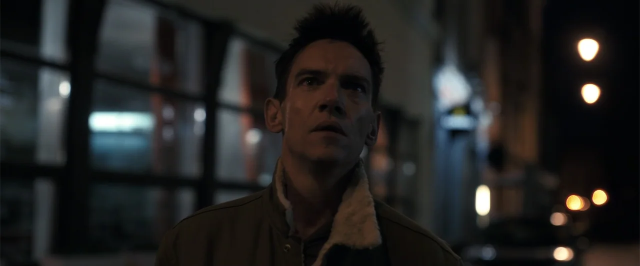 The Good Neighbor (2022) movie still: medium — A dark urban sidewalk at night with blurred streetlights and storefront windows in background. A man…