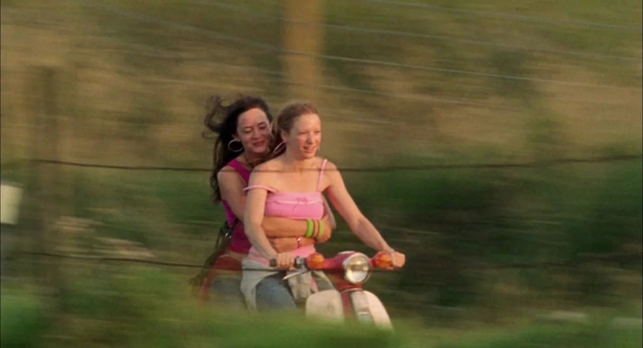 My Summer of Love movie still: medium — Two women ride a red scooter along a rural grass-lined dirt road at sunset. The background is blurred with…