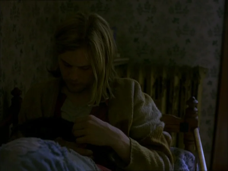 Last Days (2005) movie still: medium — A dimly lit room with patterned wallpaper features a radiator against the back wall. A young man with long…