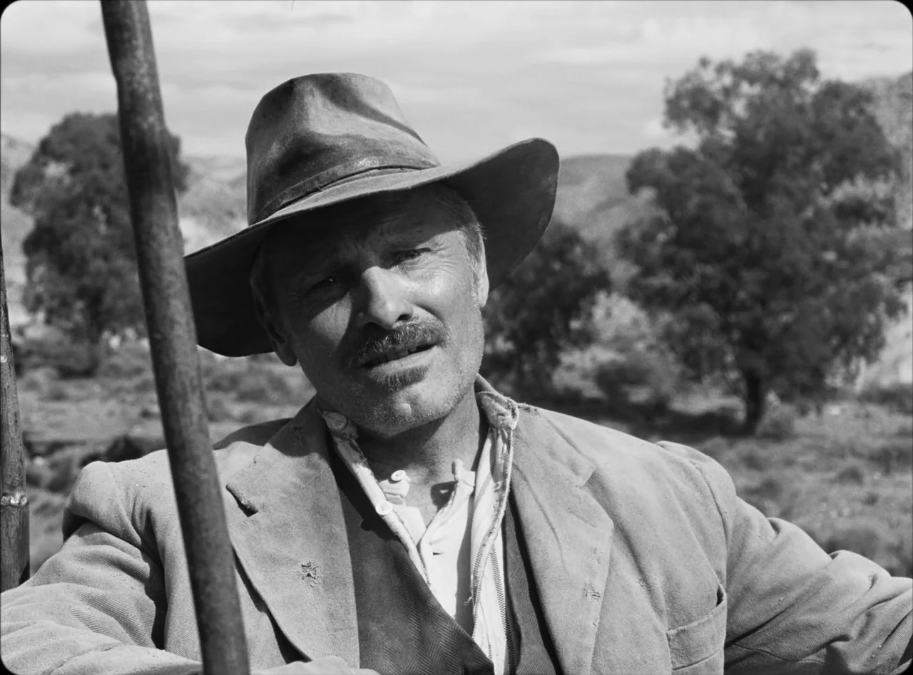 Eureka (2024) movie still: medium — An arid, open rural landscape with sparse trees and scattered shrubs behind the subject. A man wearing a…