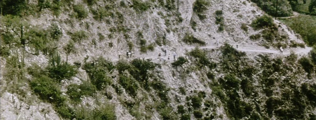Japón (2002) movie still: wide — A steep, arid mountain slope features a thin, unpaved path winding through dry scrub. A small group of people walks…