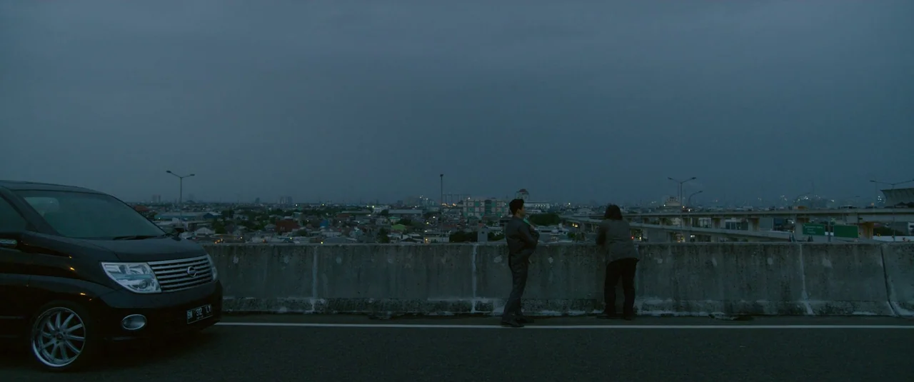 The Night Comes for Us (2018) movie still: wide — An asphalt road runs past a concrete overpass railing overlooking a sprawling city. Two men stand…