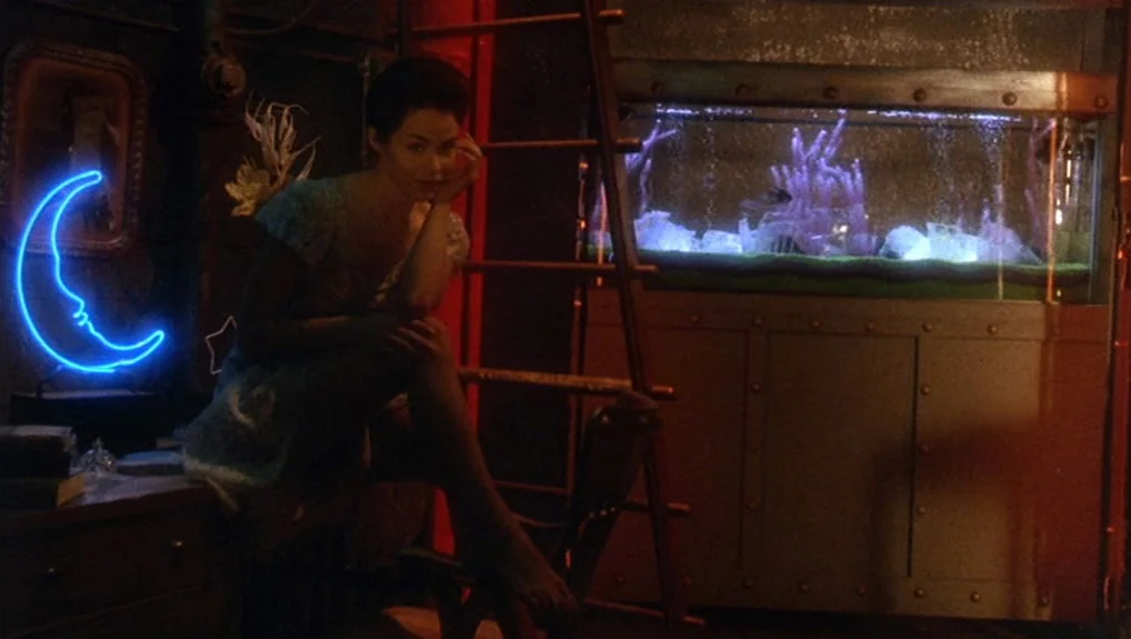 Graffiti Bridge (1990) movie still: medium — A dimly lit room contains a large aquarium and a blue neon moon sign. A woman sits on a chair in the…