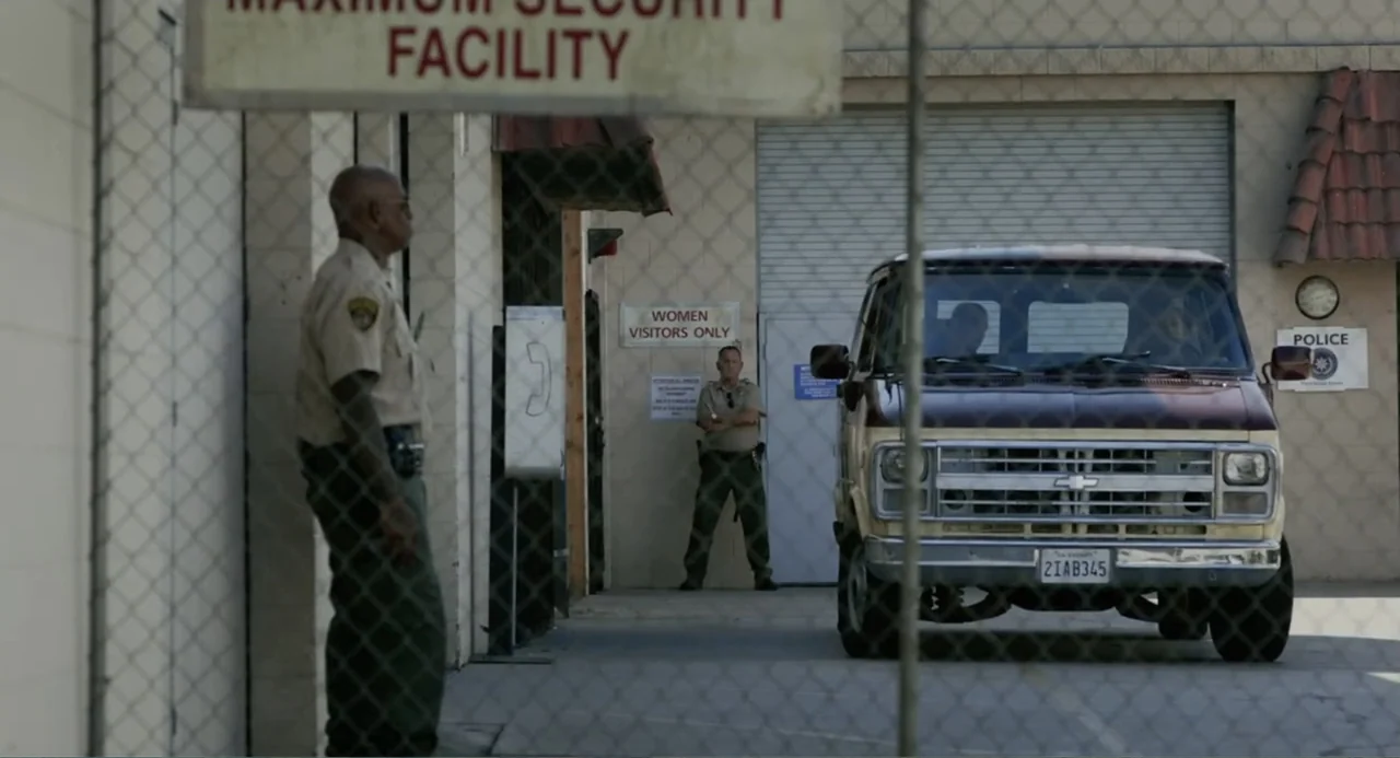 Elles (2018) movie still: wide — A secured facility entrance with a chain-link fence in the foreground. A police guard stands watch near a van…