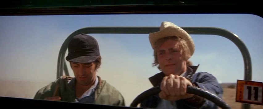 Scarecrow (1973) movie still: medium — Two men travel through a dusty landscape inside an open-top vehicle. The driver wears a cowboy hat while the…