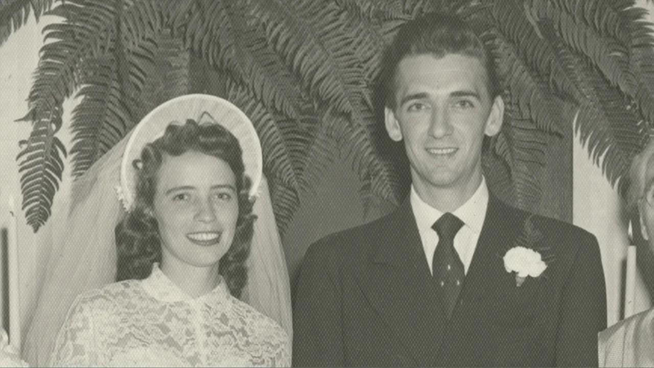 The Winding Stream (2014) movie still: medium — A wedding photograph displays a bride and groom standing before a large fern plant. The couple poses…
