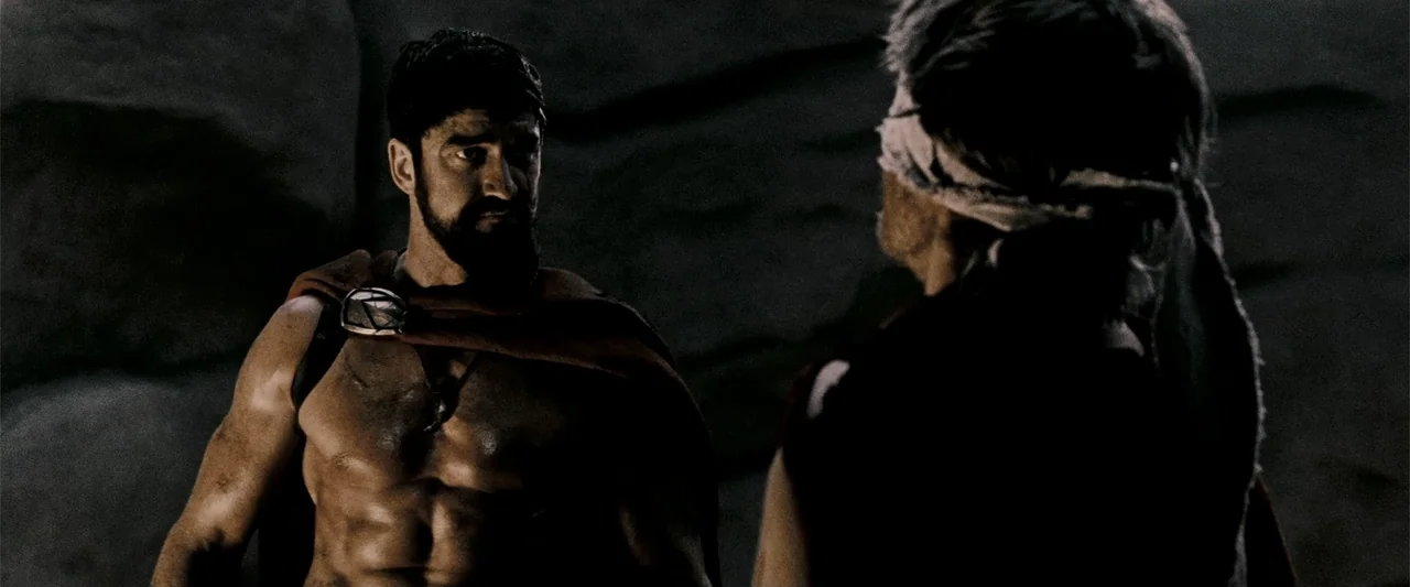 300 (2007) movie still: medium — A rocky, barren mountain pass with textured stone walls in the background. A muscular, shirtless warrior faces a…