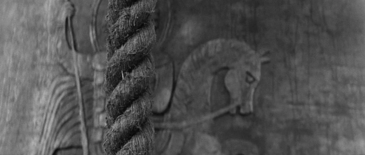 Andrei Rublev (1966) movie still: closeup — A thick fiber rope hangs vertically in front of a stone wall. The wall features a relief carving of a…