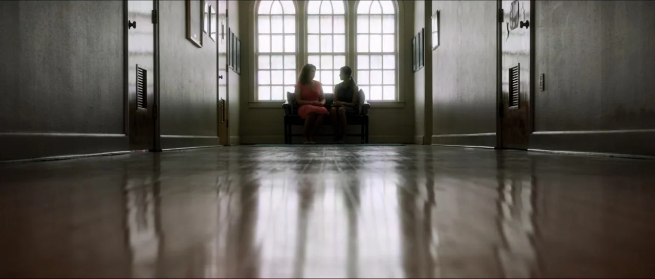 If You're Gone (2019) movie still: wide — A long hallway with polished wooden floors leads to large arched windows. Two women sit on a bench…
