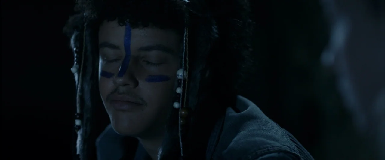 Nocturne (2020) movie still: closeup — A dark outdoor setting at night with a person wearing a decorative fur hat. The individual sits with their…