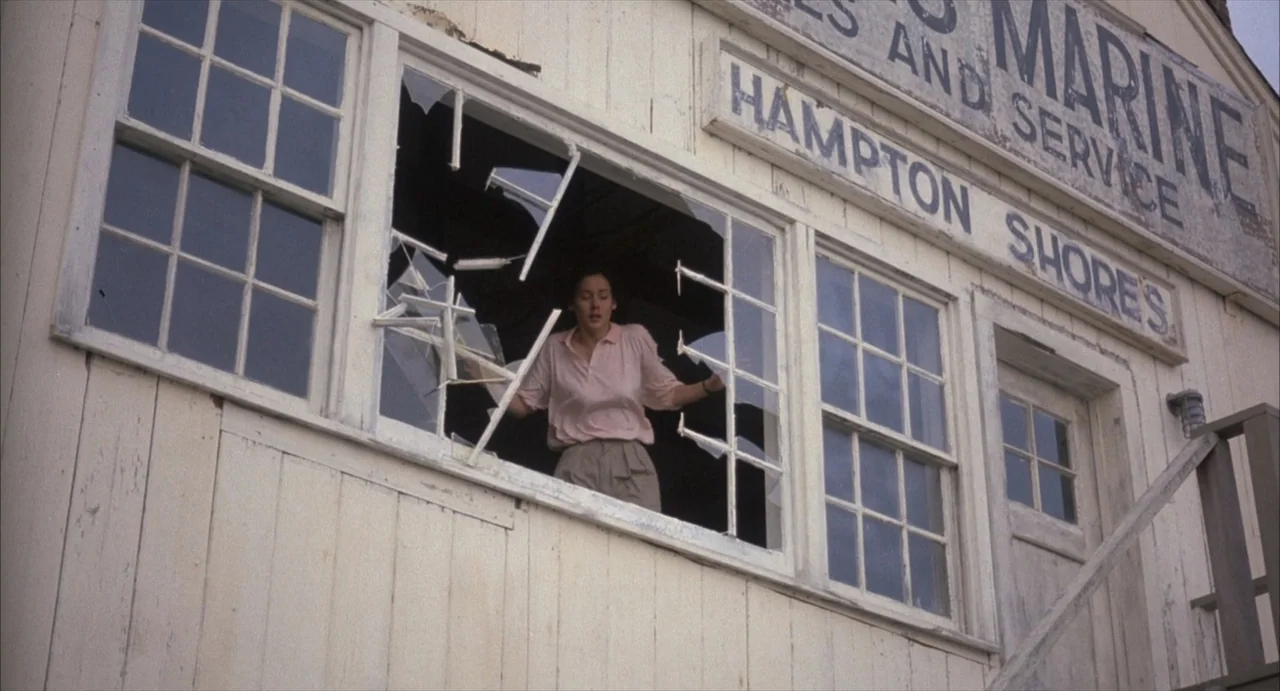 Masquerade (2023) movie still: medium — A weathered white wooden building exterior features a large broken window with jagged glass. A woman climbs…