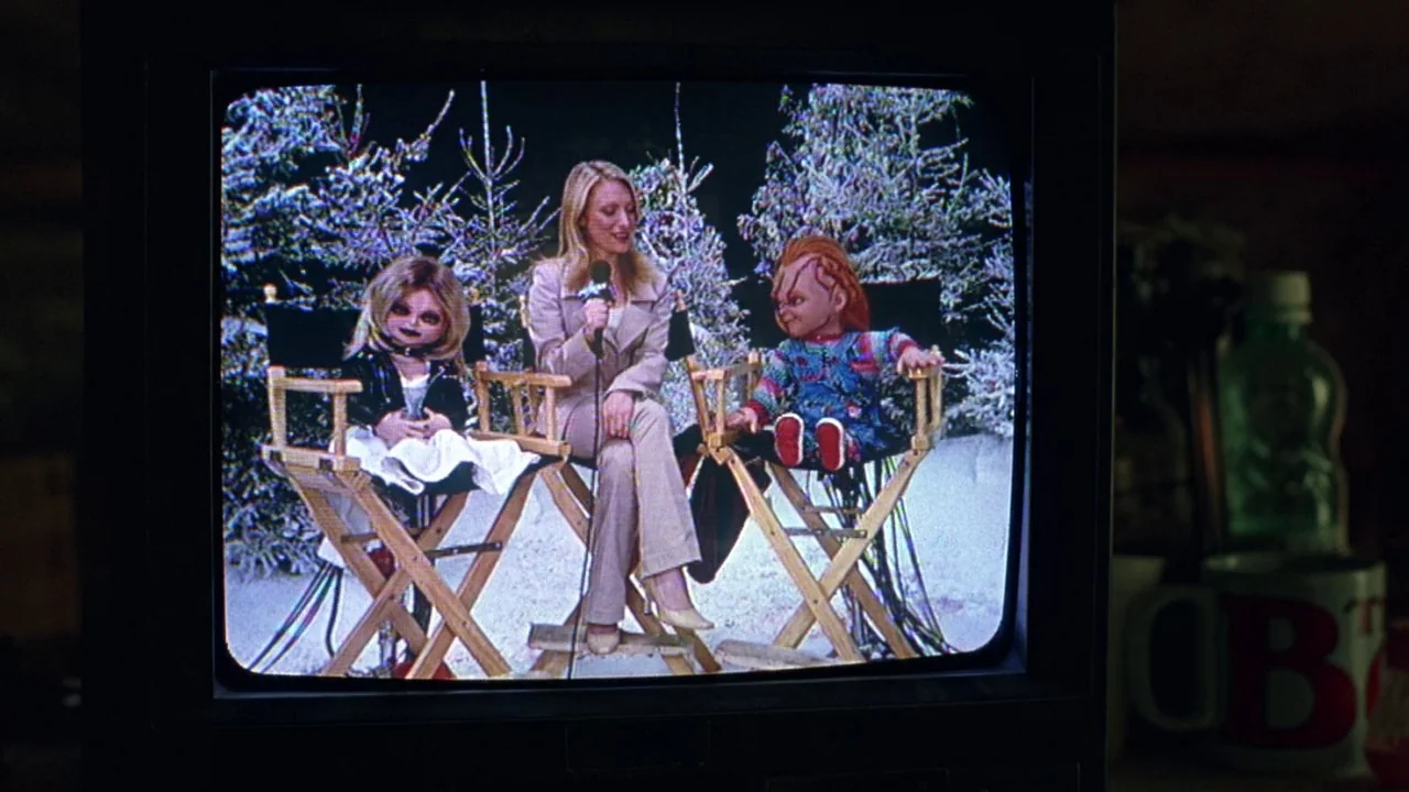 Seed of Chucky (2004) movie still: medium — A woman sits between two dolls on a television studio set with snowy decorations. The scene is broadcast…