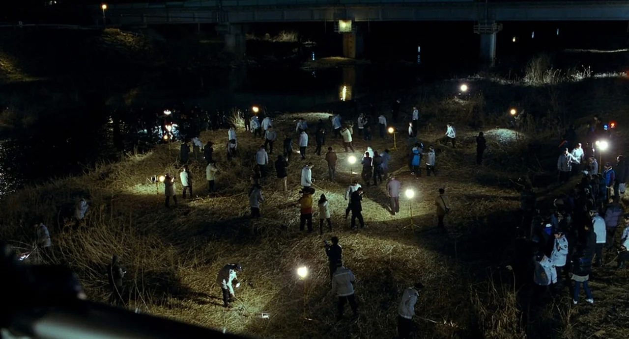 I Saw The Devil (2010) movie still: wide — A dark riverbank at night with tall, dead grass covers the uneven terrain. A large group of people scans…