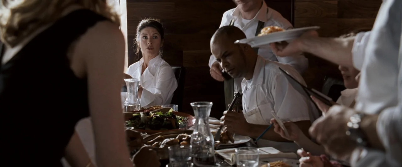 No Reservations (2007) movie still: medium — A restaurant dining table is set with food and glassware in a modern interior. A woman and a man in…