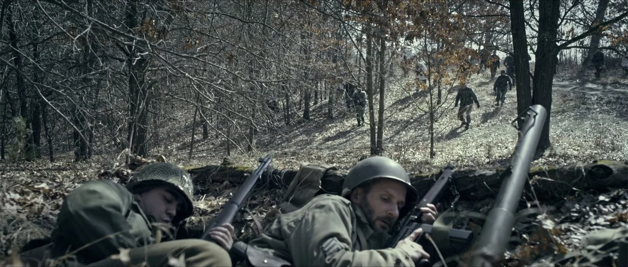 Battle of the Bulge (1965) movie still: medium — A group of soldiers is positioned in a defensive trench within a winter forest. Two soldiers lie in…