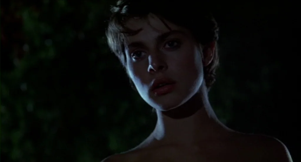 Cat People (1982) movie still: closeup — A dark night exterior features thick green foliage blurring into shadow behind a young woman. The woman…