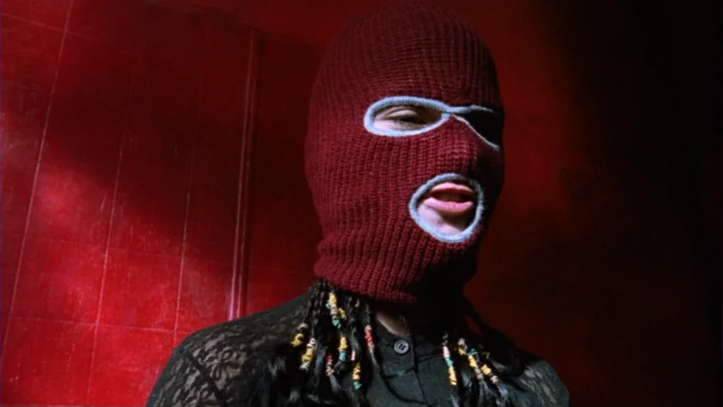Patty Hearst (1988) movie still: closeup — A dark space with vivid red tiling on the walls. A person wearing a burgundy balaclava speaks while…