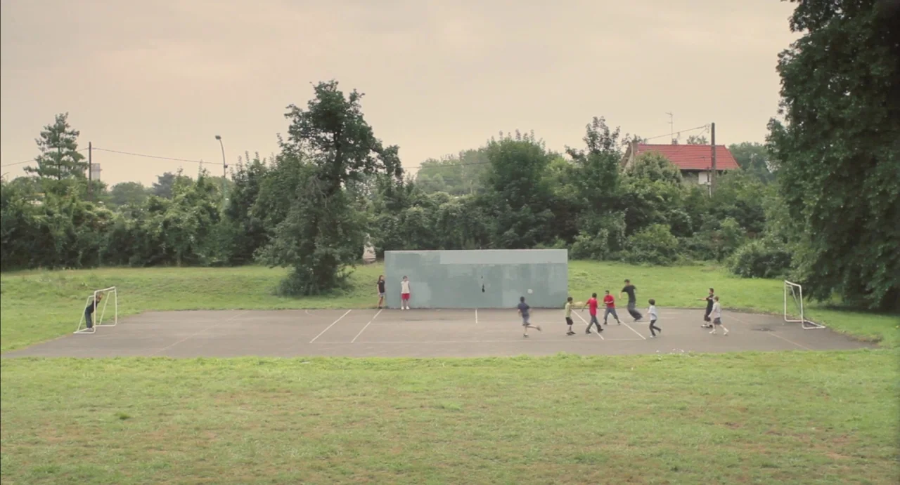 Tomboy (2011) movie still: wide — An asphalt sports court sits in the middle of a grassy, treelined outdoor area. A small group of children runs…