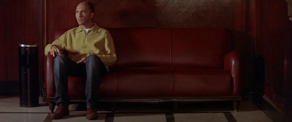 Talk To Her (2002) movie still: medium — A dimly lit room with dark wood-paneled walls and a tiled floor. A man sits alone on a large red leather…