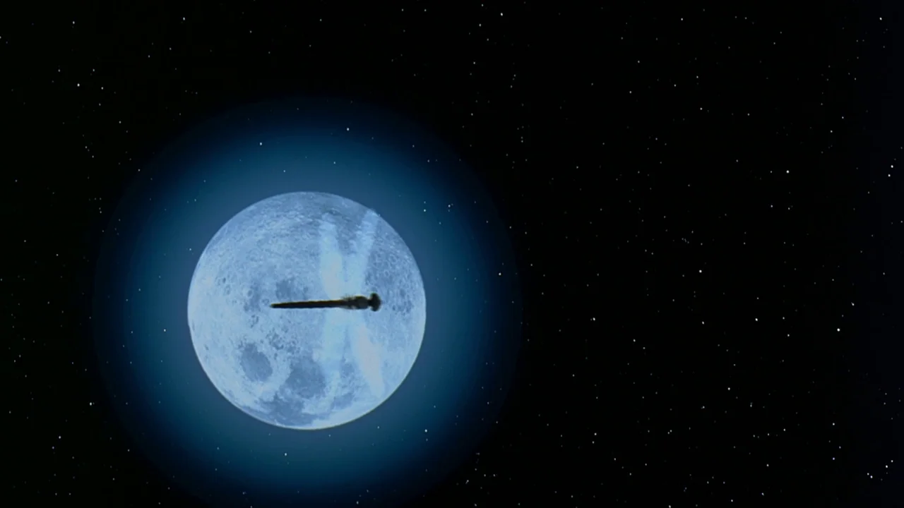 Men In Black (1997) movie still: wide — A dark, silhouetted spacecraft transits across the center of a bright, full moon. The background is an…