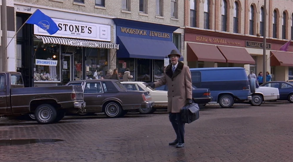 Groundhog Day (1993) movie still: fullbody — A row of storefronts lines a paved town square with several parked vehicles. A man in a tan trench coat…