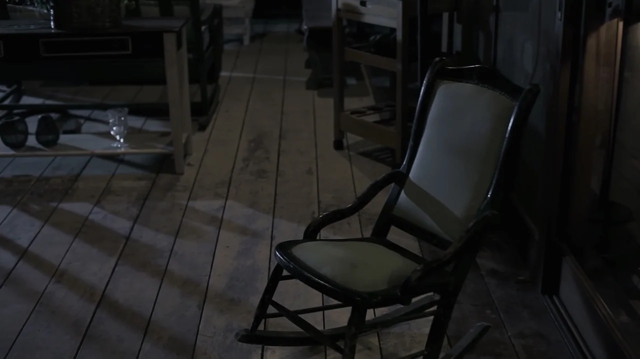 Still Life (2013) movie still: medium — A weathered wooden porch at night features a dark rocking chair with a pale cushion. The empty chair remains…