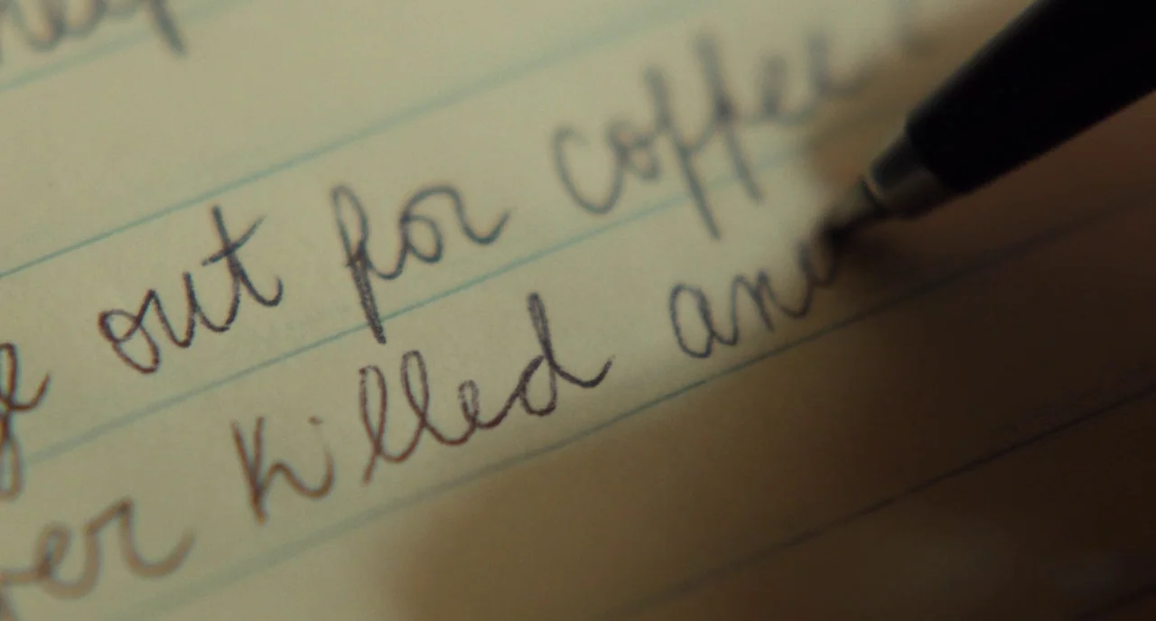 Christine (2016) movie still: closeup — A close-up view of ruled cream-colored paper with cursive handwriting in dark ink. A black ballpoint pen tip…