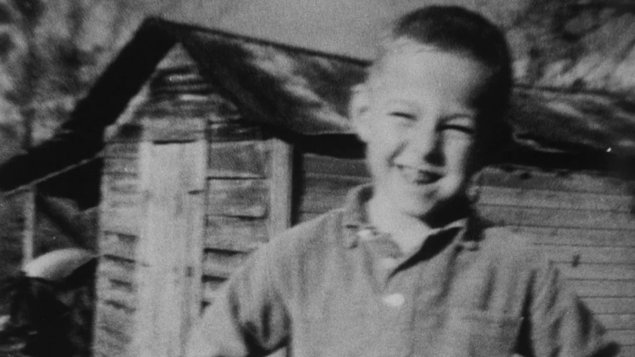 The Thin Blue Line (1988) movie still: medium — A young child stands in an outdoor area before a small wooden shed. The boy smiles directly toward…