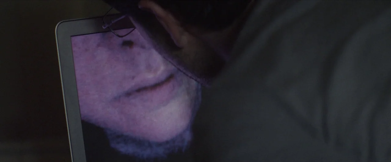 Chevalier (2015) movie still: closeup — A dark, dimly lit indoor space with low ambient light levels. A person looks down at the bright screen of a…