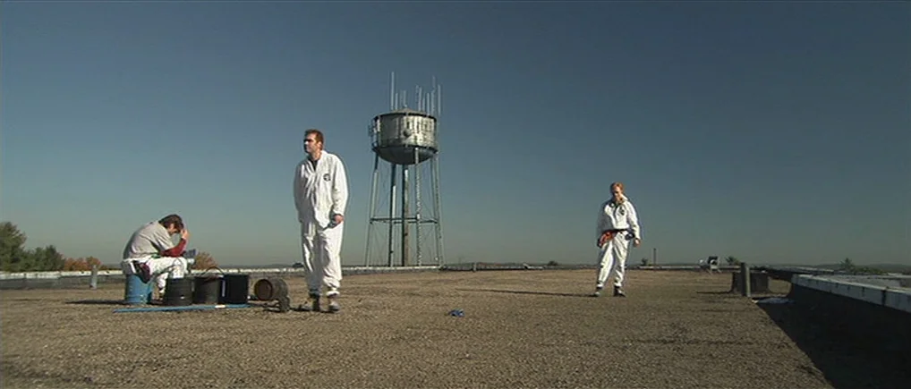 Session 9 (2001) movie still: wide — An expansive flat commercial roof features a tall metal water tower in the center. Three people dressed in…
