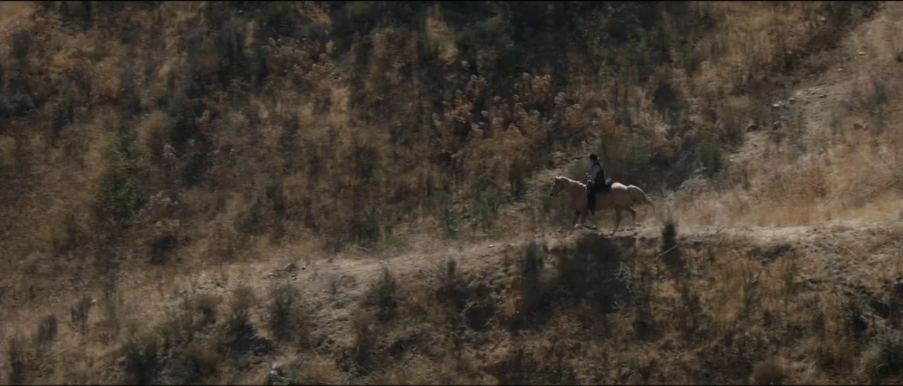 Dead for a Dollar (2022) movie still: wide — A dry, brush-covered hillside slopes steeply upward on the left side of the frame. A lone person rides…