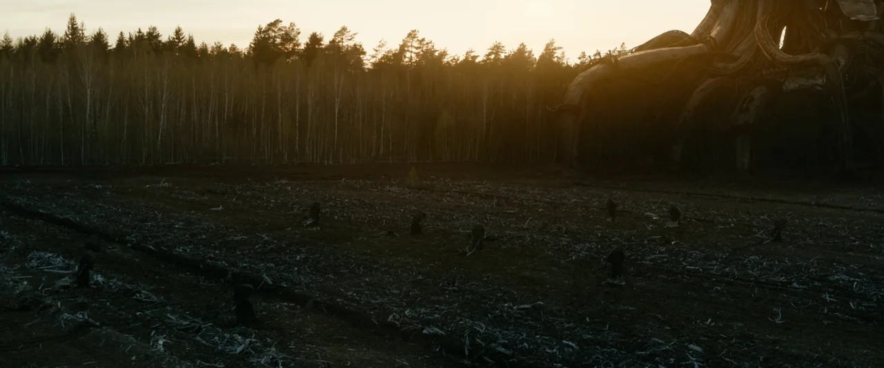 Vesper (2022) movie still: wide — A dark, cleared field filled with debris sits in front of a dense pine forest. A massive, non-terrestrial metallic…