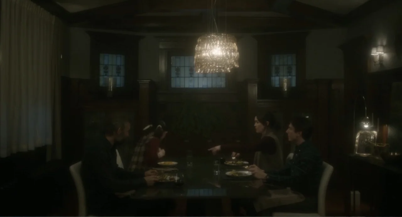 Orphan: First Kill (2022) movie still: medium — A dark dining room features wood paneling and a modern glass pendant lamp overhead. Four people are…