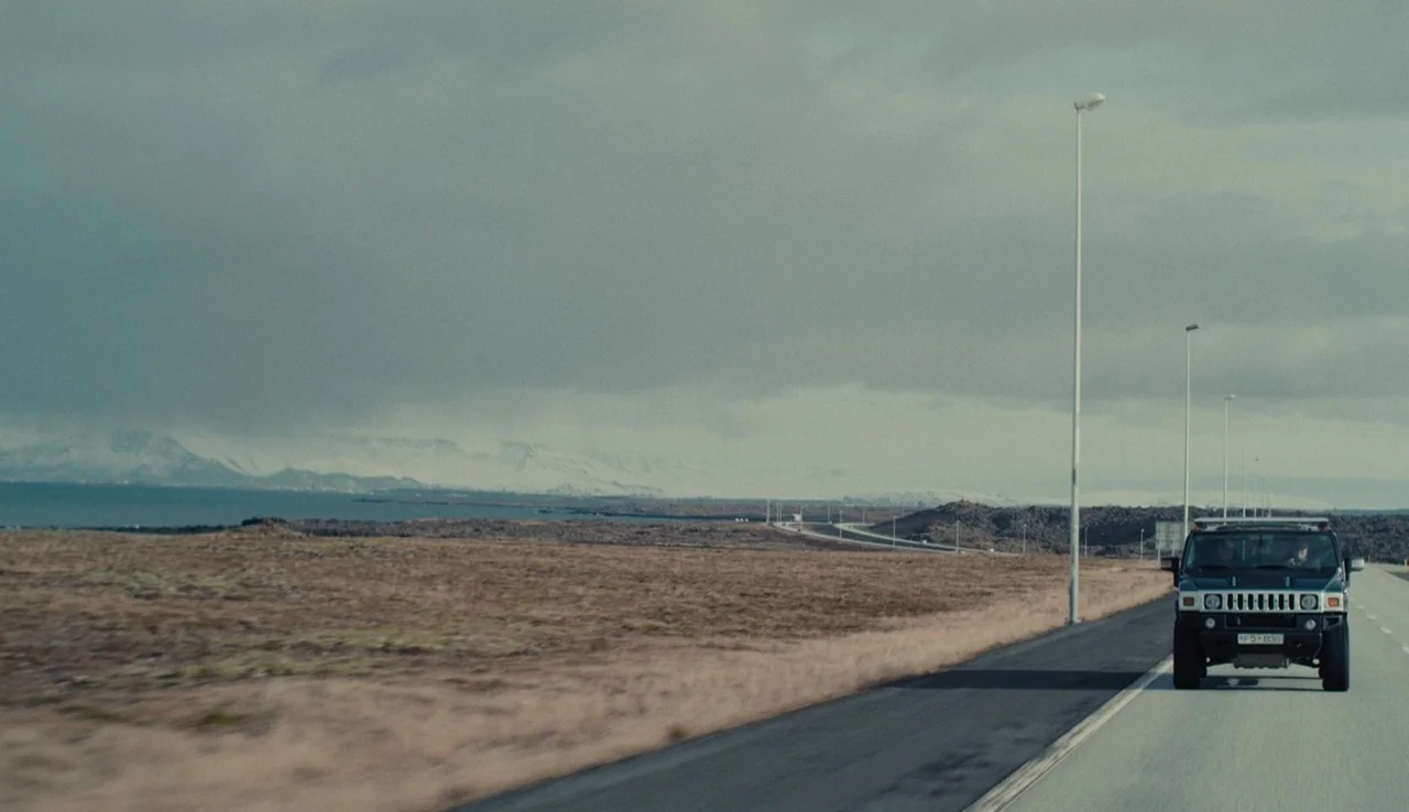Land Ho! (2014) movie still: wide — An asphalt highway stretches across a barren, flat landscape under a heavy overcast sky. A dark sport utility…
