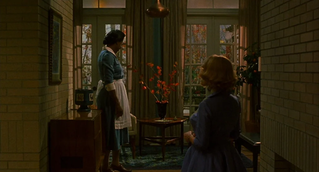 Far From Heaven (2002) movie still: medium — A mid-century home interior features light brick walls and floor-to-ceiling glass panel doors. Two…