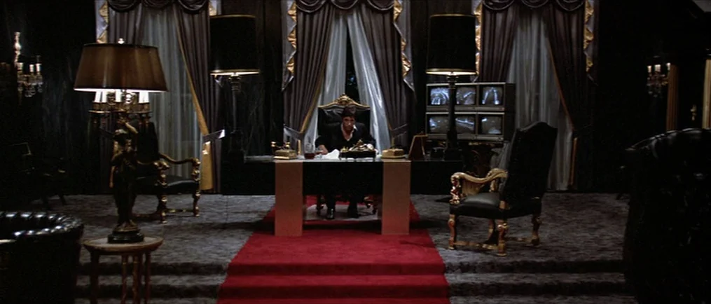 Scarface (1983) movie still: wide — A spacious dimly lit room contains heavy drapery, black carpet, and a central desk. A man in a suit sits behind…