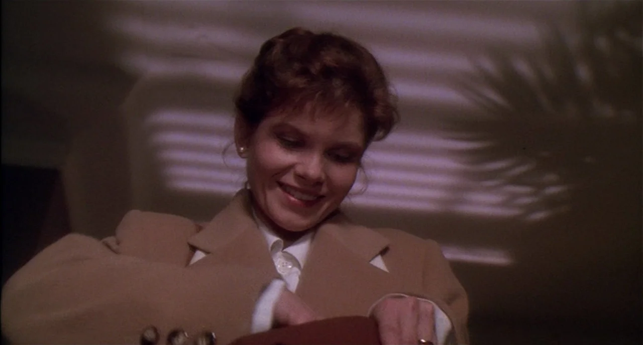 Raising Cain movie still: medium — A woman in a tan coat stands inside a dark room at night. She looks down into an open handbag with a subtle smile.