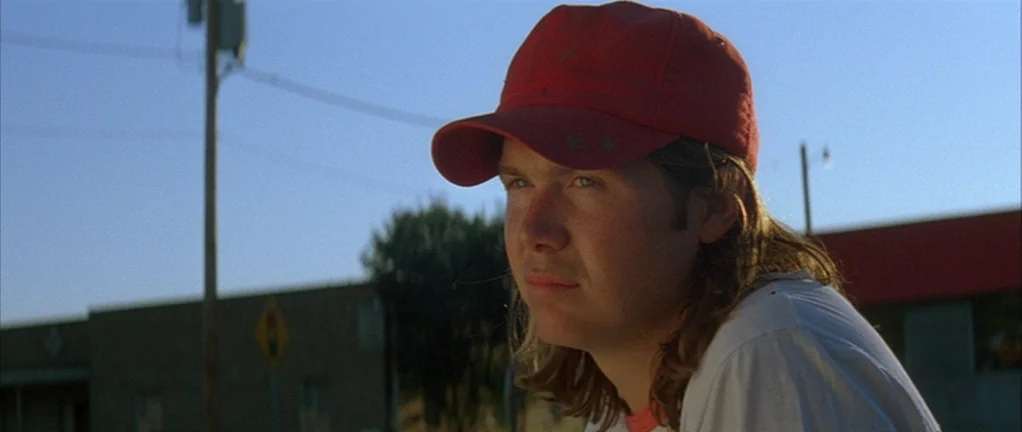 Shotgun Stories (2007) movie still: closeup — A rural setting with a clear blue sky and low-rise buildings in the distance. A teenager with long…