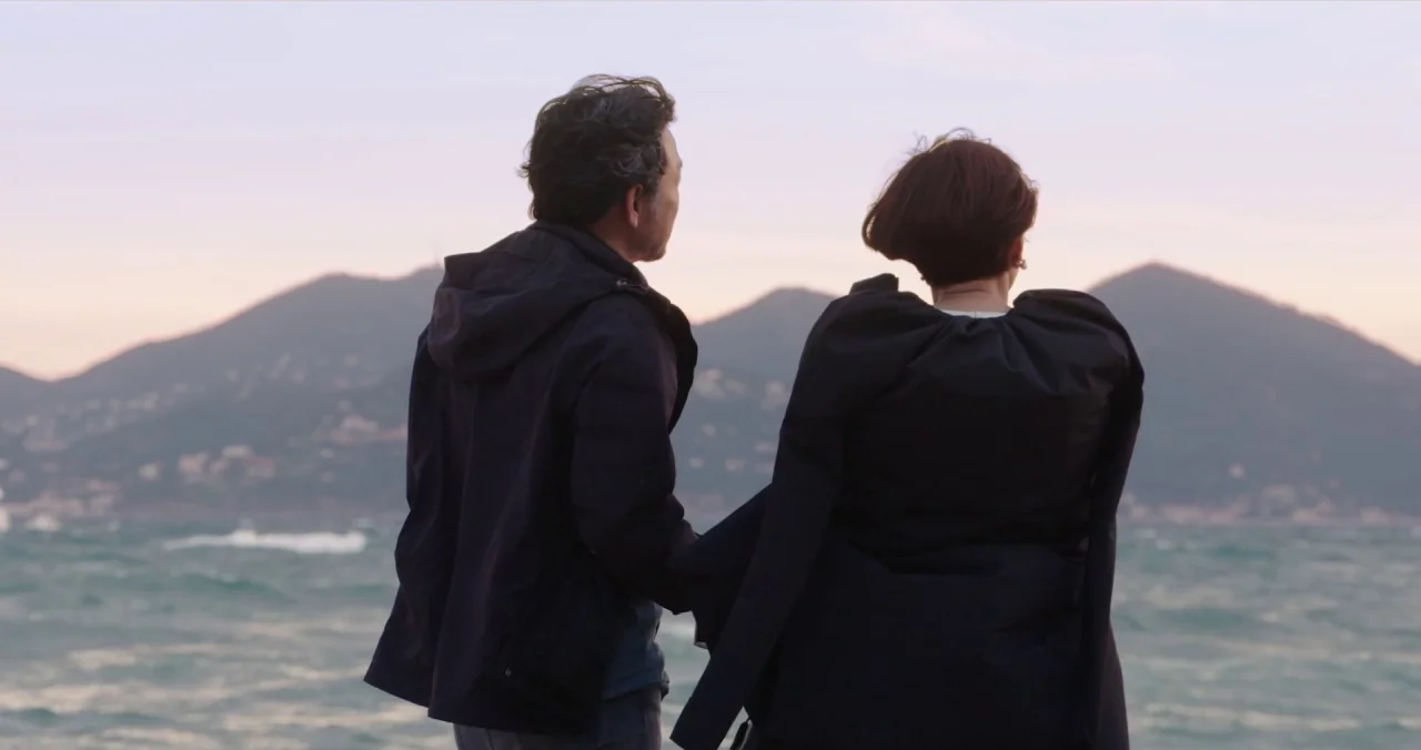 Claire’s Camera (2017) movie still: fullbody — A man and woman stand side by side before the sea at dusk. The backdrop includes rolling mountains…
