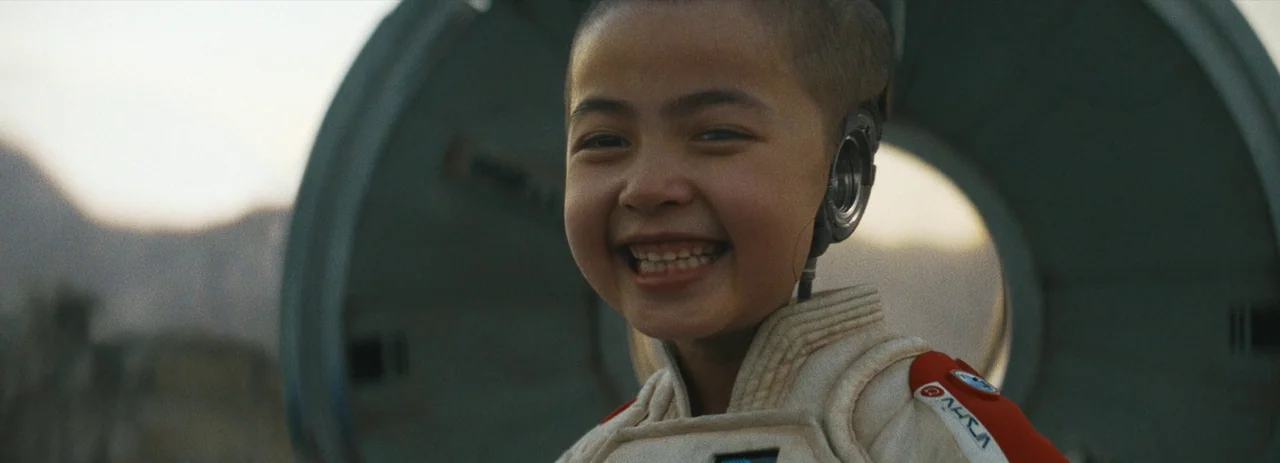 The Creator (2023) movie still: closeup — An open desert landscape serves as the backdrop for a close portrait shot. A young boy in a futuristic…