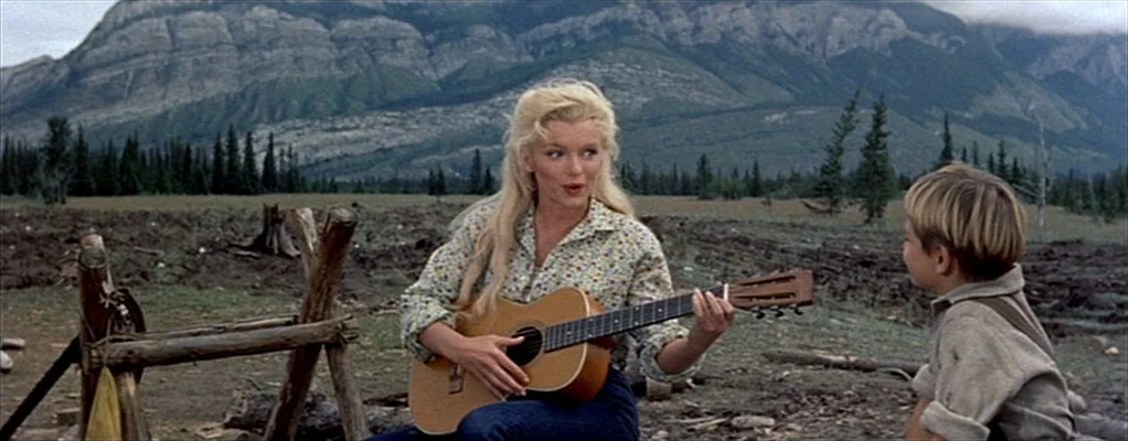 River of No Return (1954) movie still: medium — A woman sits in a clearing with mountains and a pine forest background. She plays a guitar while the…