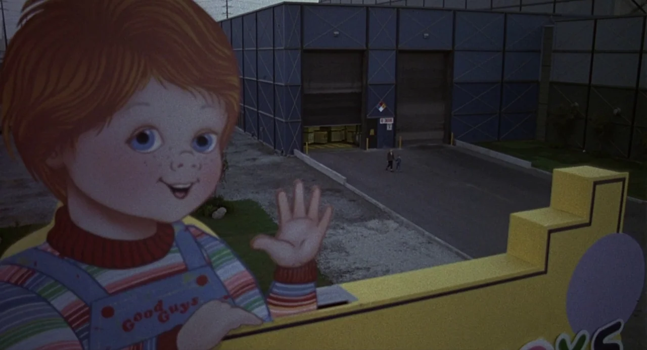 Child’s Play 2 (1990) movie still: wide — A large cardboard cutout of a Good Guys doll overlooks an industrial complex building. Two small, distant…