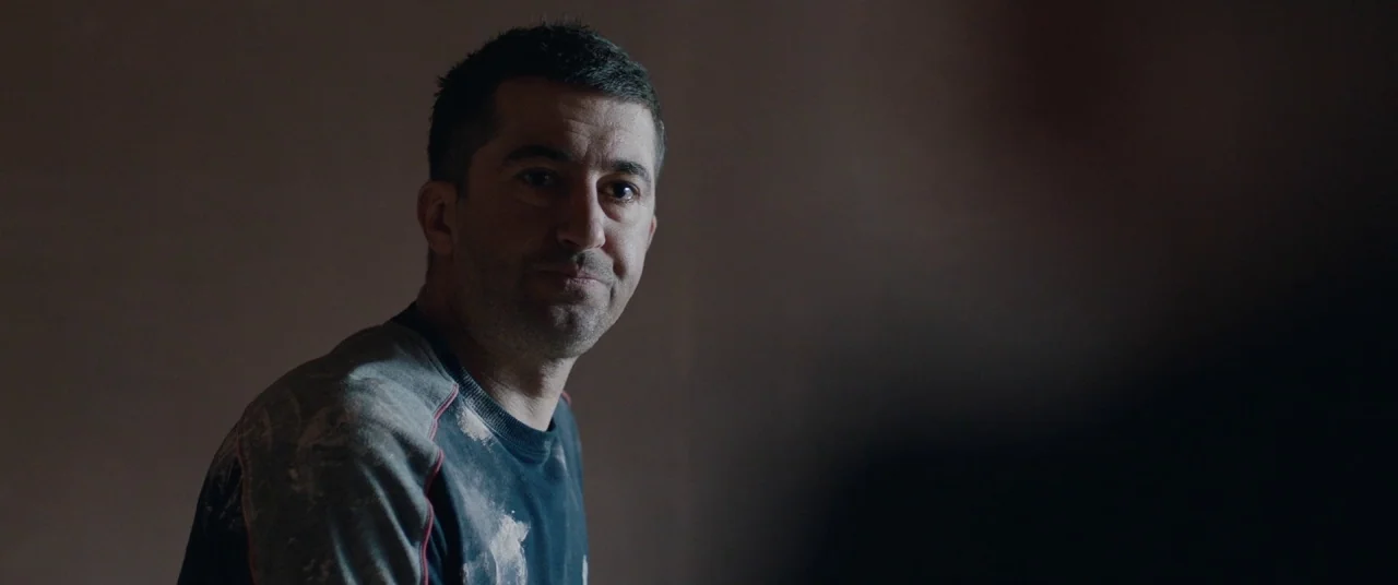 Journeyman (2017) movie still: medium — A man is positioned against a dark, undifferentiated background in a dimly lit space. He wears a stained…