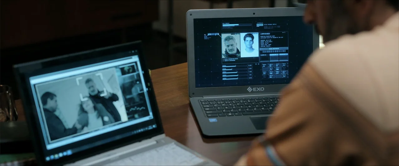 Checkmate (2015) movie still: medium — A workspace with a wooden desk holding two open laptops displaying data interfaces. A man in a patterned…
