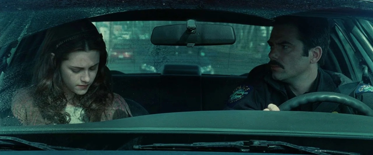 Twilight (2008) movie still: medium — A man drives a rainy police cruiser with a young woman in the passenger seat. The exterior world is visible…