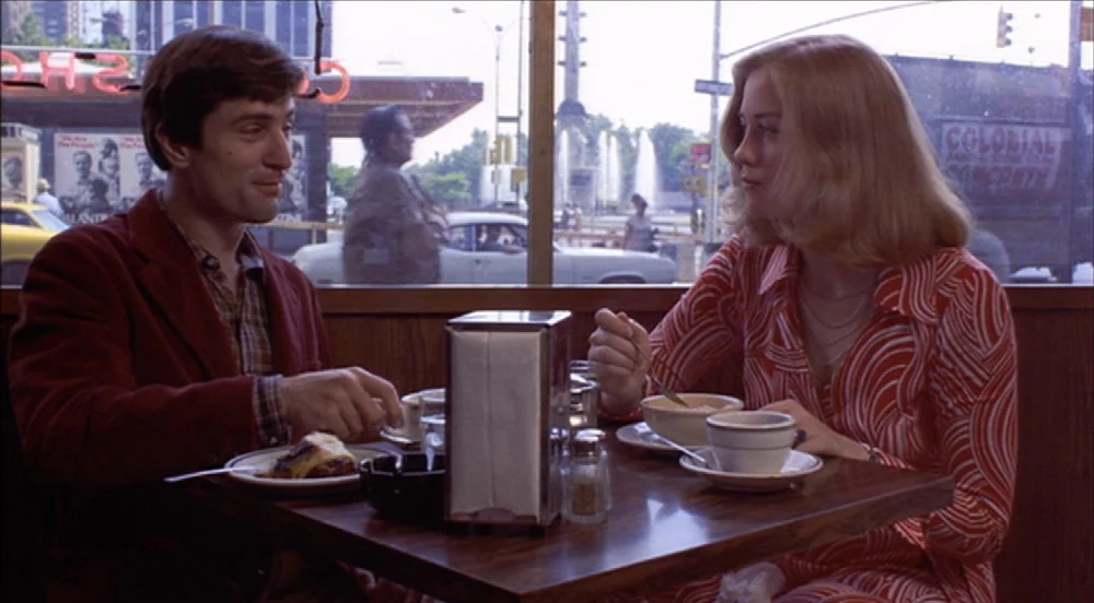 Taxi Driver (1976) movie still: medium — A 1970s diner booth with wood paneled walls overlooks a busy city street scene. A man and a woman sit…